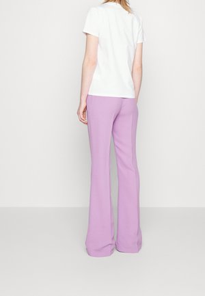 Persoon in een witte korte mouw shirt en hooggetailleerde lila wijde broek, staande met de rug naar de camera op een witte achtergrond.