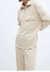 Parte superior beige con cremallera y cuello, con dos bolsillos frontales, textura suave y dobladillo redondeado, combinada con pantalones beige a juego.