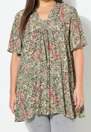 Femme portant un haut ample à manches courtes avec un motif floral abstrait vert, noir et orange et un pantalon gris, debout sur fond blanc.