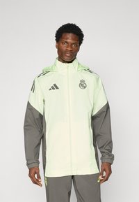 Lys grøn og grå sportsjakke med lynlås, hætte og elastiske manchetter. Har adidas striber og et logo på brystet.