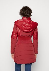 Abrigo acolchado rojo con capucha y hombros de cuero texturizado, cintura acanalada y tejido suave. Presenta costuras en relieve horizontales.