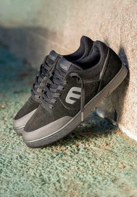 Une paire de chaussures de skate noires et grises avec des lacets, arborant un audacieux logo "E", placée contre un mur texturé sur un sol vert brut.