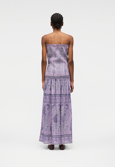Femme aux cheveux tressés portant une robe maxi violette à motifs cachemire sans bretelles, debout de dos sur un fond blanc.