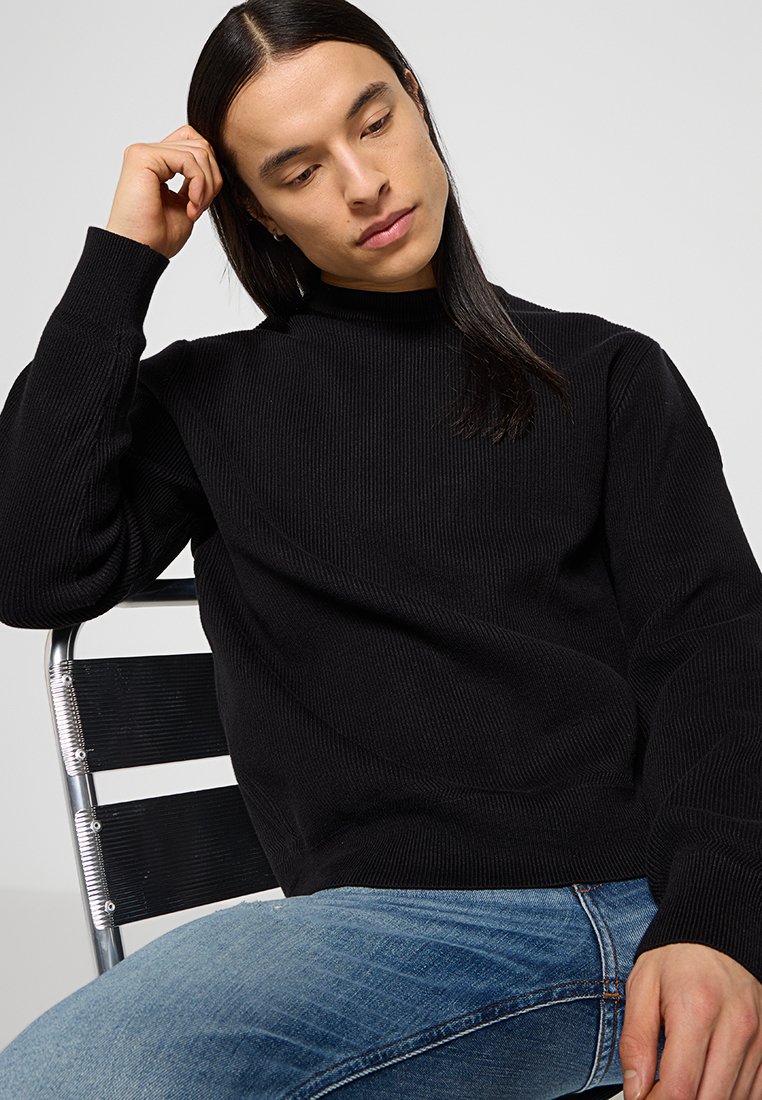 Maglione nero a coste con colletto alto, indossato con jeans blu chiaro. Modella seduta su una sedia a strisce nere, con lo sguardo rivolto verso il basso.