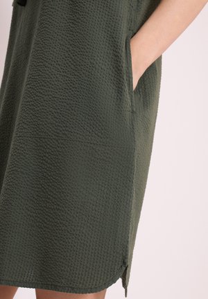 Main dans la poche d'une robe texturée vert foncé jusqu’au genou avec une petite fente latérale sur fond neutre.