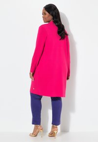 Cardigan long fuchsia à coupe droite et à manches longues, associé à un pantalon bleu foncé et des sandales à talons dorées, avec un bouton d'accent doré.