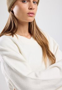 Pull blanc texturé avec un col en V et des manches surdimensionnées. Porté avec un bonnet en tricot brun, arborant de longs cheveux bruns et des bijoux minimalistes.