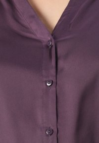 Detailní záběr na fialovou košili na knoflíky se třemi viditelnými knoflíky a částí výstřihu, ukazující hladkou texturu látky.