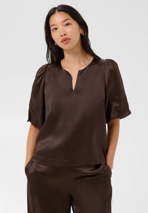 Femme aux cheveux raides et foncés portant un haut en satin marron à manches courtes et un pantalon assorti, debout les mains dans les poches sur fond gris.