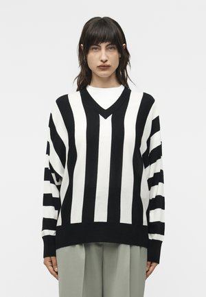 Középhosszú sötét hajú nő fekete-fehér, függőlegesen csíkozott pulóvert és világos színű nadrágot visel, egyszerű háttér előtt.
