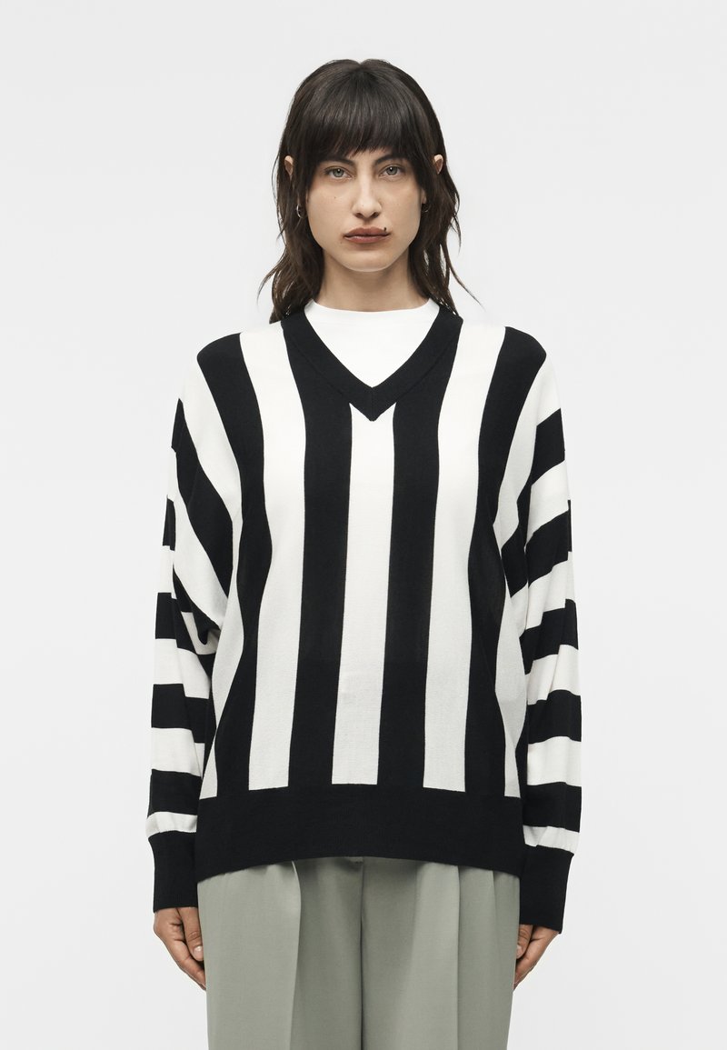 Γυναίκα με μεσαίου μήκους σκοτεινά μαλλιά που φοράει ένα ασπρόμαυρο πουλόβερ με κατακόρυφες ρίγες και ανοιχτόχρωμο παντελόνι, μπροστά σε απλό φόντο.