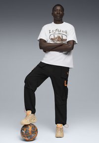 Hvid grafisk t-shirt med orange og grå design, sorte cargo-bukser med orange accenter og beige sportssko. Står på en mønstret basketball.