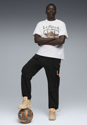 Hvid grafisk t-shirt med orange og grå design, sorte cargo-bukser med orange accenter og beige sportssko. Står på en mønstret basketball.