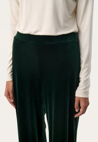 Pantaloni in velluto verde scuro con una texture liscia e una cintura elasticizzata ampia, abbinati a una maglietta a maniche lunghe color crema.