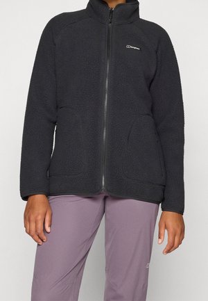 Frau trägt eine schwarze Fleecejacke mit Reißverschluss und Taschen sowie mauvefarbene Hose, gezeigt vom Hals bis zur Mitte des Oberschenkels vor einfarbigem Hintergrund.