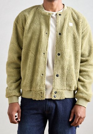 Mann trägt eine hellgrüne Fleecejacke mit Druckknöpfen über einem weißen Hemd und dunkelblauen Jeans, steht vor einem einfarbigen Hintergrund.