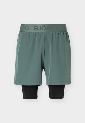 Zelené sportovní šortky s černými kompresními legínami připevněnými pod nimi a elastickým páskem s logem značky.