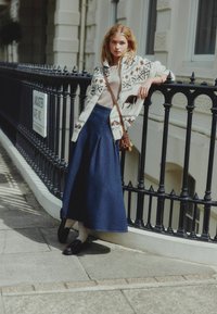 Knit cardigan with white base, brown patterns, and deer motifs, paired with a dark denim skirt and black loafers, against a wrought iron railing.