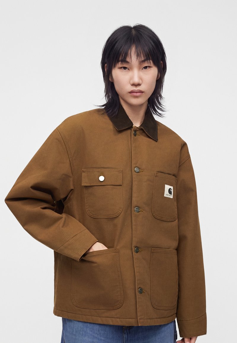Person with black hair wears a brown button-up jacket with four pockets, one showing a rectangular white Carhartt logo patch.