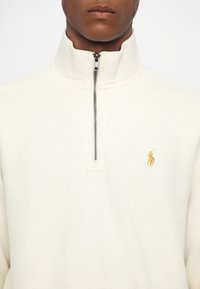 Mężczyzna ubrany w kremowy sweter z suwakiem do ćwiartki i wysokim kołnierzem oraz małym złotym haftowanym logo gracza polo na piersi.