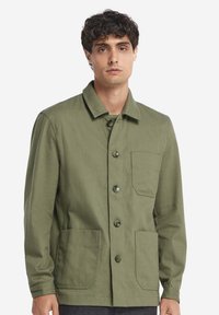 Olivgrön arbetsjacka i slitstarkt tyg, med krage, frontknappar, två påsydda fickor och en bröstficka.