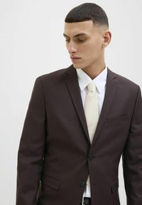 Giacca marrone con due bottoni e una tasca singola, abbinata a una camicia bianca e una cravatta crema chiara. Capelli corti e strutturati.
