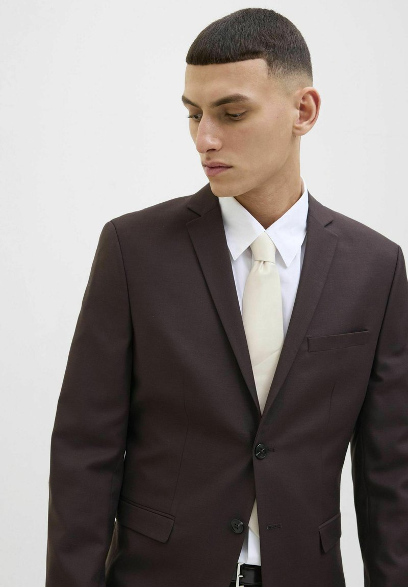 Giacca marrone con due bottoni e una tasca singola, abbinata a una camicia bianca e una cravatta crema chiara. Capelli corti e strutturati.