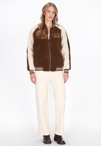 Brun og creme fleecejakke med lynlås foran, ribstrikkede manchetter og broderet tekst. Sammen med cremefarvede bukser og brune støvler.