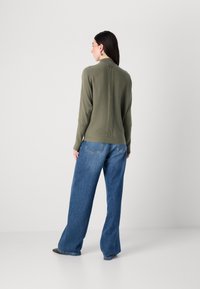 Eine Frau steht mit dem Rücken zur Kamera, trägt einen olivgrünen Pullover und weite blaue Jeans, die einen minimalistischen Modestil zur Schau stellen.