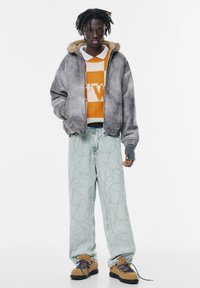 Jachetă bomber gri cu glugă pufoasă, pulover portocalie cu model, pantaloni din denim bleu deschis cu cusături contrastante și pantofi bej cu șireturi.