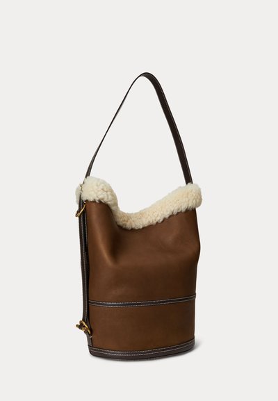 Brun skinnbucketväska med foder i fårskinn, bred axelrem, två horisontella dekorlinjer och gulddetaljer. Mjuk textur med strukturerad form.
