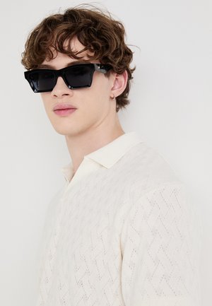 Fekete, téglalap alakú napszemüveg vastag kerettel, amit egy krémszínű, textúrált pólót viselő személy hord, amelynek galléros nyaka van.