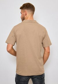 Polo shirt à manches courtes de couleur tan avec un tissu texturé, un col et un ourlet droit ; porté avec un jean gris foncé, montrant la vue arrière.