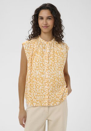 Kobieta o kręconych ciemnych włosach, ubranapłaszcza w żółto-białą wzorzystą bluzkę bez rękawów i jasnobeżowe spodnie, stoi na tle jednolitego tła.