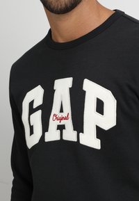 Sudadera negra de tejido suave con grandes letras blancas "GAP" y texto rojo "Original" en un estilo colegial.