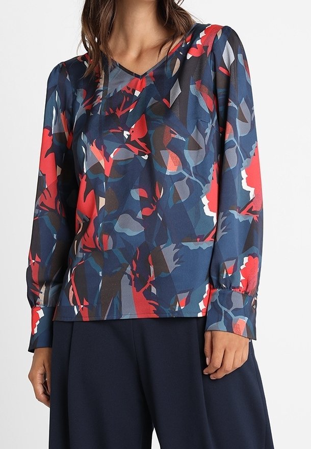 Femme portant un chemisier à manches longues avec un motif floral abstrait rouge, marine et gris, associé à un pantalon ample bleu marine foncé.