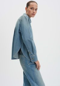 Giacca in denim con vestibilità morbida e colletto a punta, caratterizzata da un colore azzurro chiaro, grandi bottoni e un design con schiena spaccata per una migliore ventilazione.