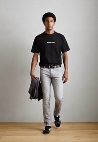Zwarte T-shirt met witte tekst, grijze spijkerbroek, grijze trui in de hand en zwarte sportschoenen. Eenvoudig ontwerp, fitted vorm en neutrale kleuren.