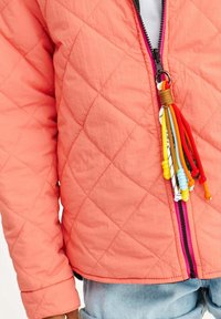 Korálový prošívaný kabát se zipem vpředu. Má barevné třásně na táhle zipu a jemný lesk v látce.