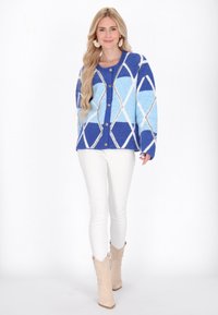 Světle modrý a modrý cardigan s vzorem argyle, s knoflíky, měkkou texturou a oversized střihem, kombinovaný s bílými kalhotami a béžovými kotníkovými botami.