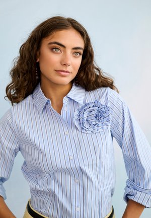 Mladá žena s vlnitými hnedými vlasmi má na sebe modro-biele pruhované tričko s látkovým kvetom na hrudi, pozerá sa mierne do strany.