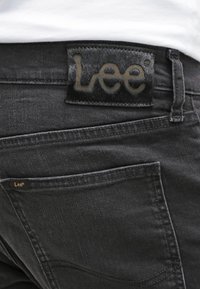 Svarta denimjeans med en texturerad, svart läderlapp med "Lee"-logotyp. Inkluderar standardfickor och synliga sömdetaljer.