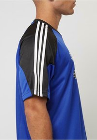 Blå sportströja med svarta ärmar, prydd med tre vita ränder på varje axel, tillverkad av ventilerande tyg, med rundad halsringning.