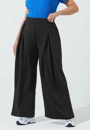 Donna che indossa pantaloni neri a gamba larga con pieghe, camicia blu a maniche corte e bottoni, e scarpe da ginnastica bianche su sfondo neutro.