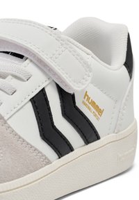 Sneaker bianca e nera con tomaia in materiale sintetico, inserti in suede, tre strisce nere, cinturino in velcro e suola bianca testurizzata. Presenza di branding dorato.