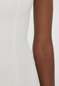 Gros plan d'un bras à peau brune à côté d'un vêtement blanc avec des lignes de couture visibles sur un fond clair.