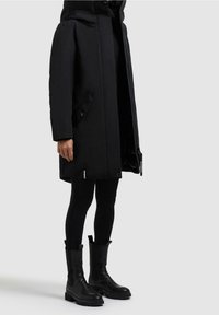 Personne portant un long manteau noir à capuche, des leggings noirs et des bottes noires montantes jusqu'à mi-mollet, debout devant un fond uni.