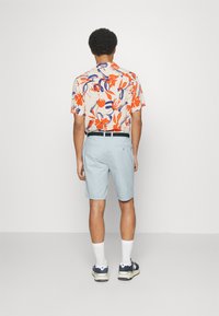 Chemise à manches courtes florale en beige avec des motifs orange et bleu, associée à un short bleu clair et des chaussettes blanches avec des chaussures foncées.