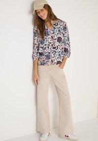 Femme portant une casquette beige, un chemisier à manches longues à motifs floraux, un pantalon large beige et des baskets blanches, debout contre un mur blanc en souriant.