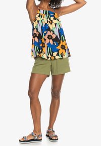 Personne portant un haut sans manches coloré à fleurs, un short vert olive et des sandales à fleurs, se tenant avec les bras levés devant un fond blanc.